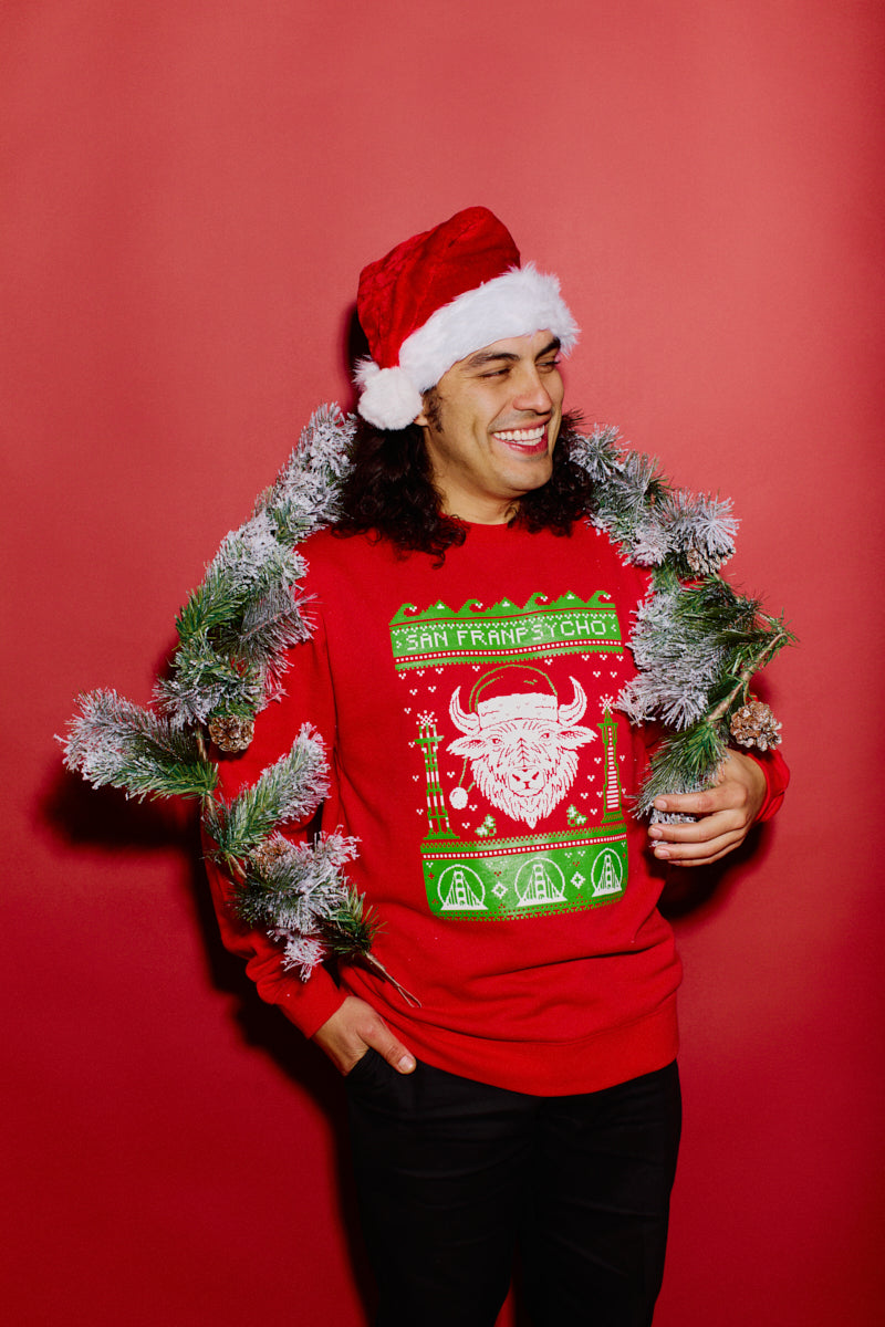 A smiling person wearing an SFP Holiday Crewneck and a Santa hat holds a holiday garland against a red background, ready to spread cheer.