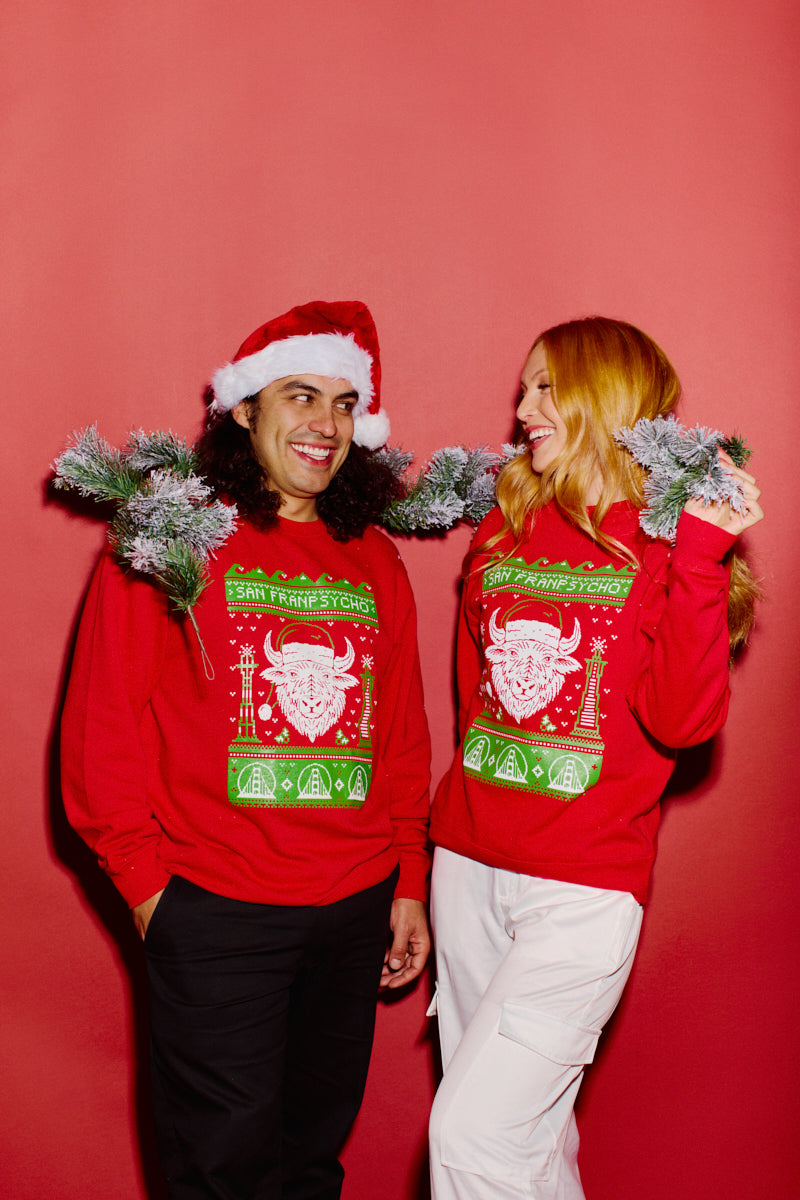 Two people wearing the SFP Holiday Crewneck and Santa hats are smiling against a red background, with pine garlands draped around their necks.