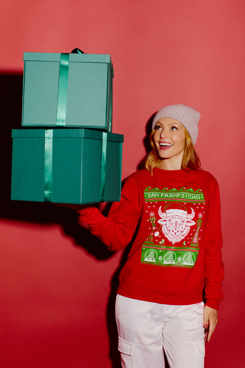 A person wearing an SFP Holiday Crewneck and a beanie holds two green gift boxes against a vibrant red background.