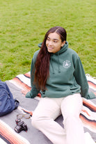 A woman in a hand-printed green Battle Cat Pullover Hoodie relaxes on a striped blanket in the grass, with her camera and bag nearby.