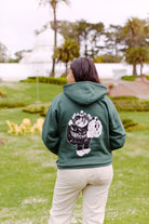 In a park with palm trees and a white building, someone wears the green Battle Cat Pullover Hoodie, showcasing a hand-printed graphic on the back.