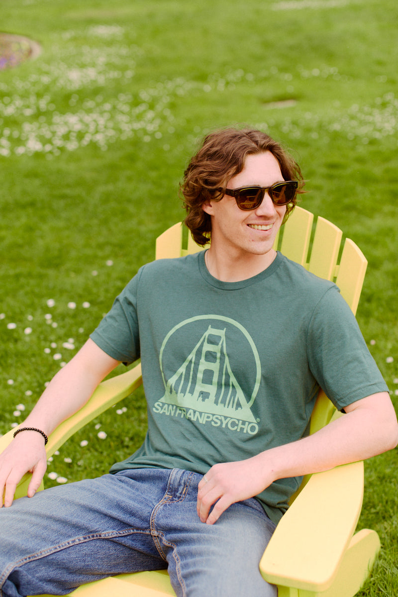 A man in sunglasses sits on a yellow chair outdoors, wearing jeans and the Green Logo Tee, known for its comfort.