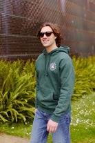 A person wearing a green Battle Cat Pullover Hoodie and sunglasses smiles while standing outdoors near ferns and a metal structure.