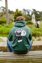 Someone wearing a green Battle Cat Pullover Hoodie, made from a poly/cotton blend and showcasing a cartoon design, sits on a bench in the park.