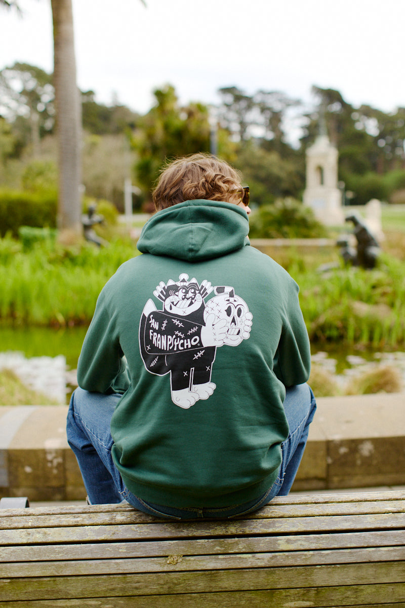 Someone wearing a green Battle Cat Pullover Hoodie, made from a poly/cotton blend and showcasing a cartoon design, sits on a bench in the park.