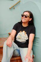 A person in stylish sunglasses and a Womens Cat House Tee - Black sits on outdoor stairs against a light blue wall, smiling. This 100% cotton womens cut T-shirt adds casual elegance to the sunny day scene.