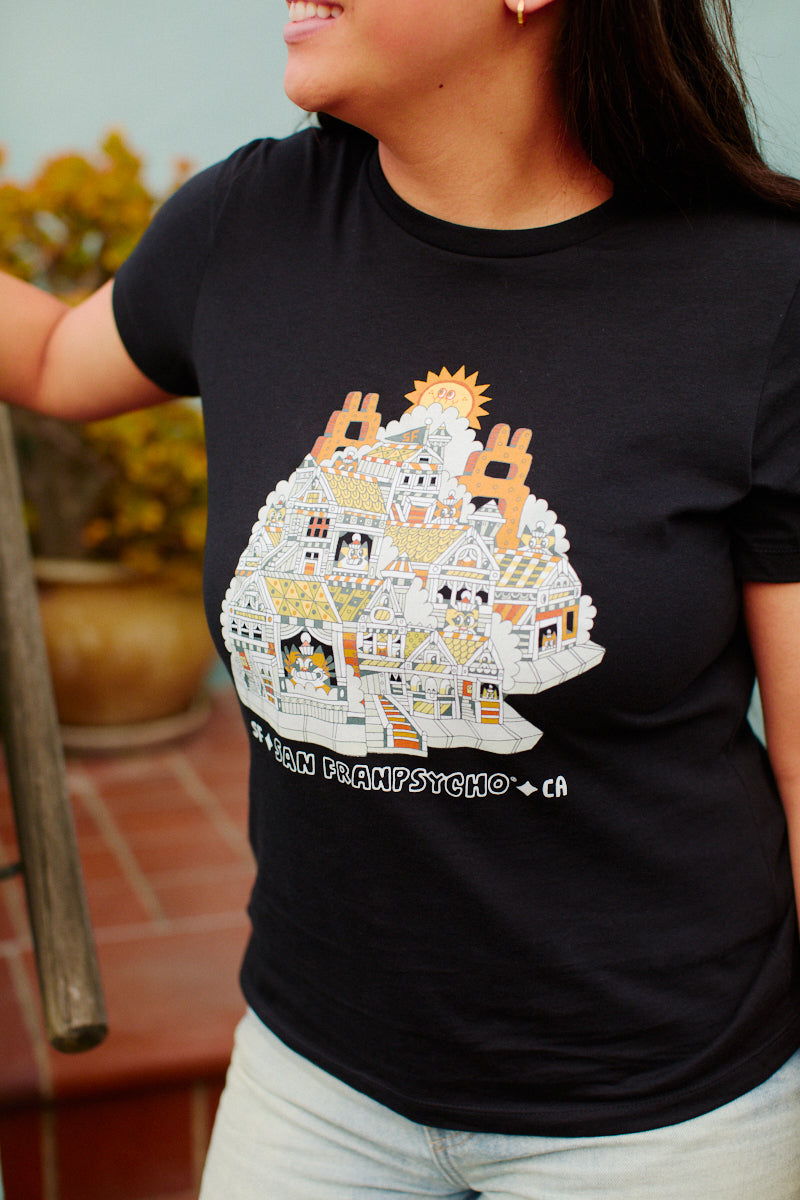 A person is standing outdoors wearing a Womens Cat House Tee - Black, featuring a San Francisco cartoon house design. The tee is designed for women and made from 100% cotton.