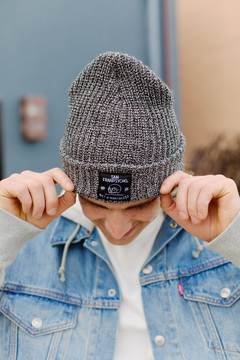 Wearing the Zebra Lumberjack Beanie w/ Wet & Wreckless, a person in a denim jacket looks down and adjusts their beanie, showcasing its bold style and unique logo detail.