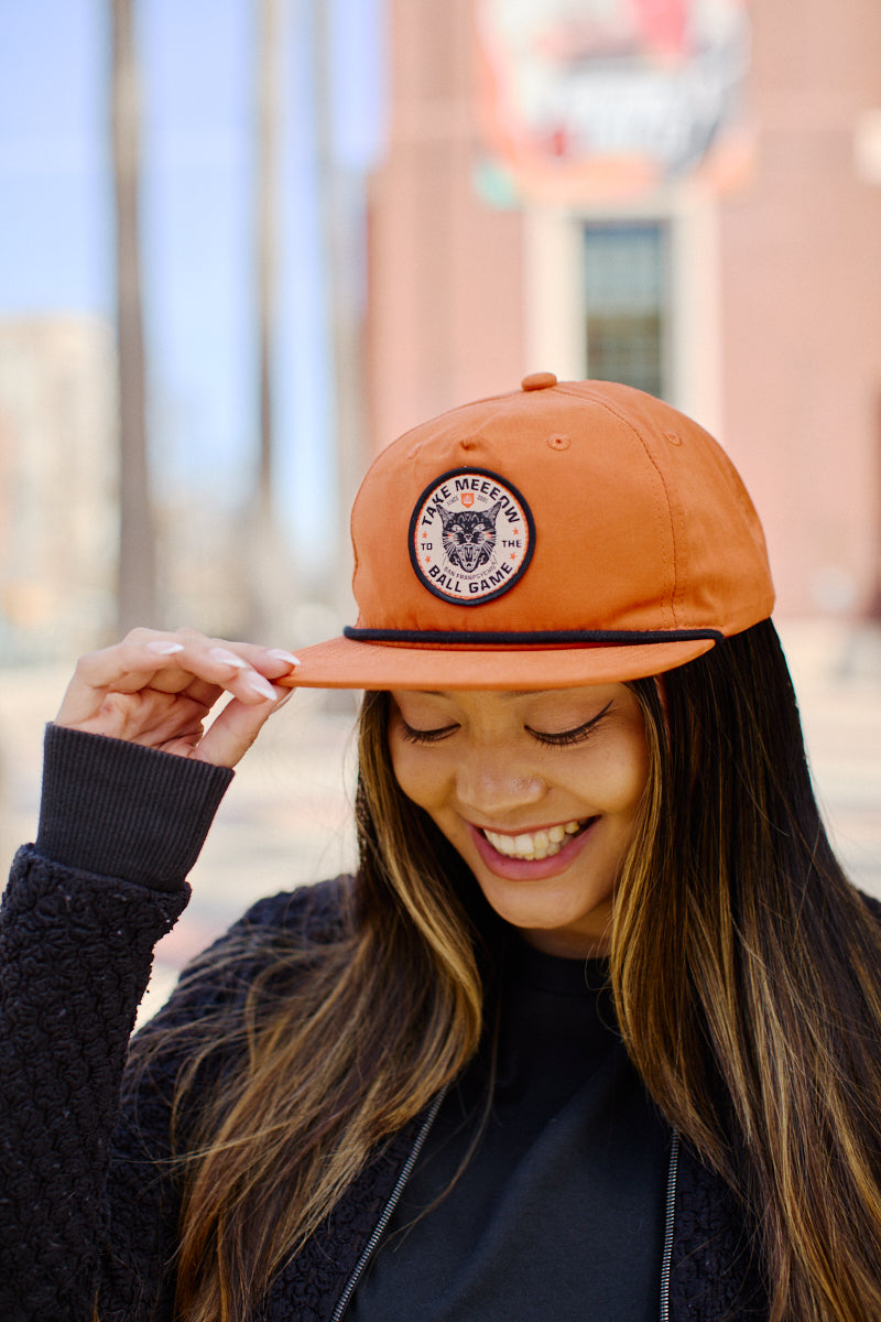 A smiling woman in an orange Take Meeeow Patched Hat basks in sunshine with a lively outdoor backdrop, embracing the baseball season spirit.