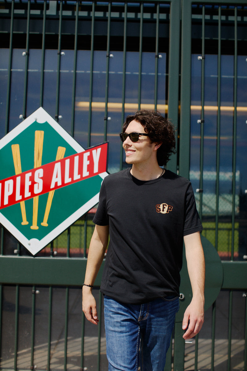 A man wearing a Psychos Logo Tee and sunglasses walks past a fence with a baseball-themed sign, embodying black & orange pride. His hand-printed San Francisco shirt perfectly complements his casual vibe.