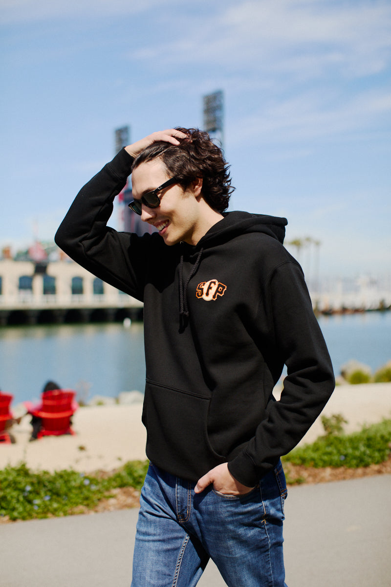 A person in a sleek Psychos Logo Pullover Hoodie walks along the San Francisco waterfront, their sunglasses adding an air of mystery as they pass buildings under a clear sky.