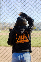 A person wearing the Black & Orange Logo Pullover stands before a chain-link fence, gazing at the baseball field.