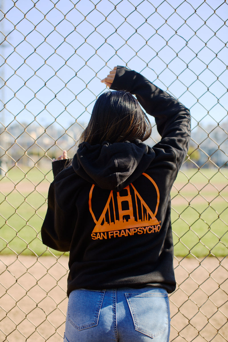 A person wearing the Black & Orange Logo Pullover stands before a chain-link fence, gazing at the baseball field.