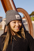 A woman in a Zebra Lumberjack Beanie w/ Natural & Black Logo and black sweater smiles outdoors on a sunny day.