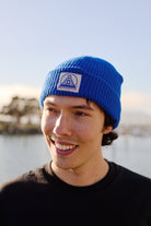 Person happily wearing a Royal Blue Waffle Beanie with Natural/Blue Logo, standing outdoors by the water.