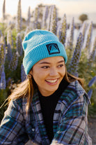 In a field of tall purple flowers, a person beams as they stand outdoors wearing a plaid shirt and the Cyan Waffle Beanie w/ Black/Blue Logo.