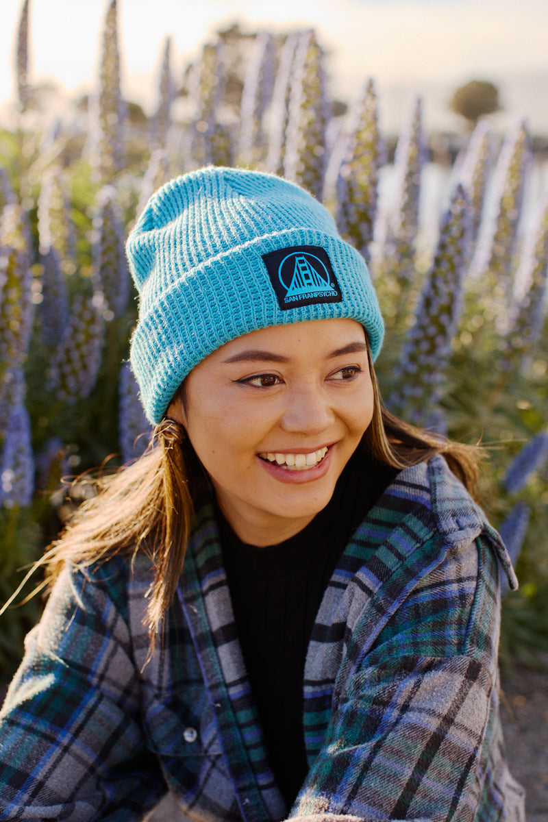In a field of tall purple flowers, a person beams as they stand outdoors wearing a plaid shirt and the Cyan Waffle Beanie w/ Black/Blue Logo.