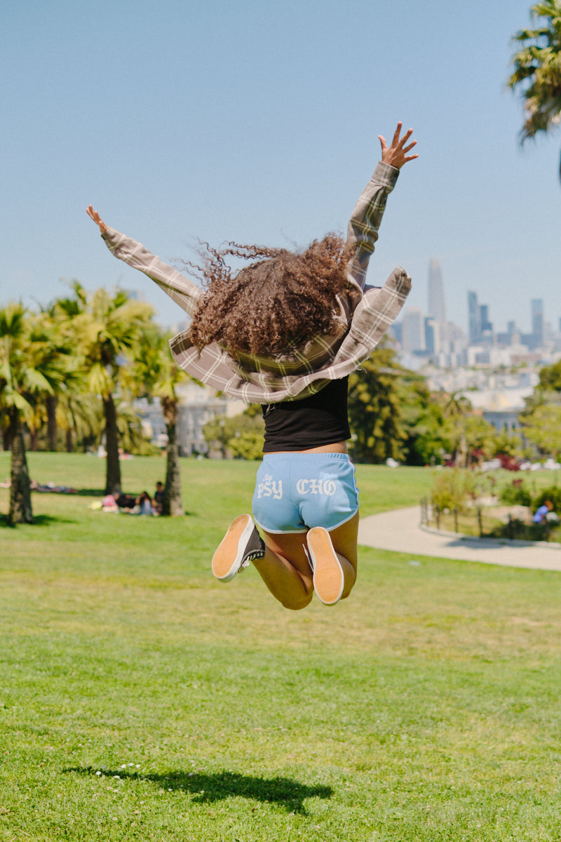 Leaping joyfully in a park with the city skyline and palm trees behind, this person wears Stay Psycho Booty Shorts - Blue, perfect for a sunny day.