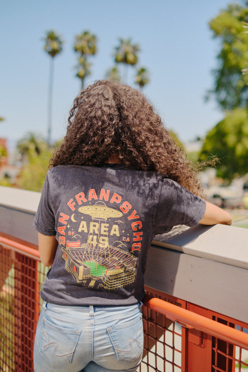 A person with curly hair wears the Area 49 Tee, leaning on a red outdoor railing with palm trees in the background.