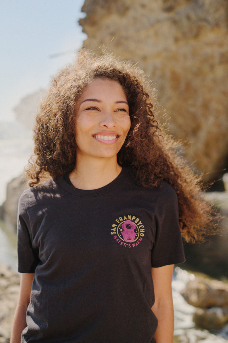 A woman with curly hair smiles outdoors near rocks and water, wearing the Psycho Siren Tee—hand-printed on soft Airlume cotton.