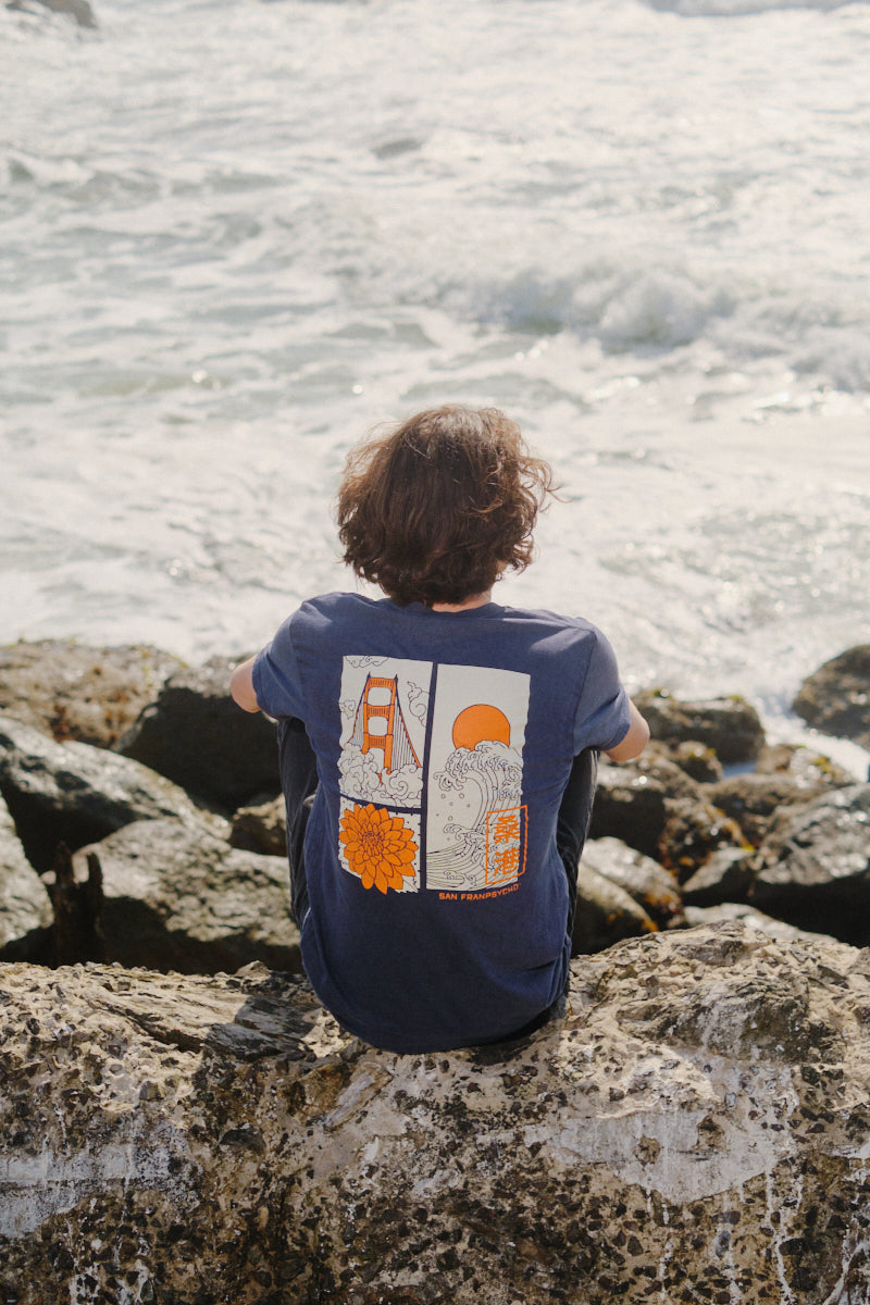 A person with curly hair sits on oceanfront rocks, wearing the blue Sunset Dahlia Tee featuring vibrant, Japanese art-inspired graphics on the back.