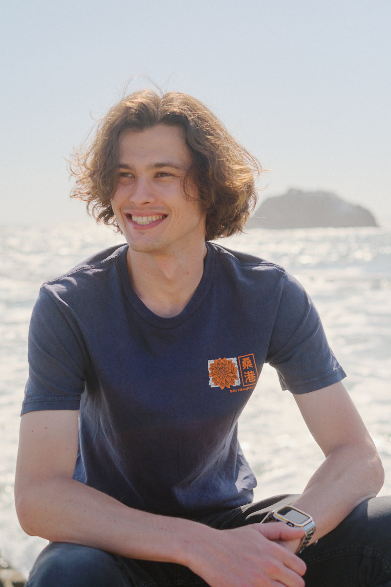 A young man with wavy hair smiles by the ocean in a Sunset Dahlia Tee, inspired by Japanese art, sunlight glowing behind him.