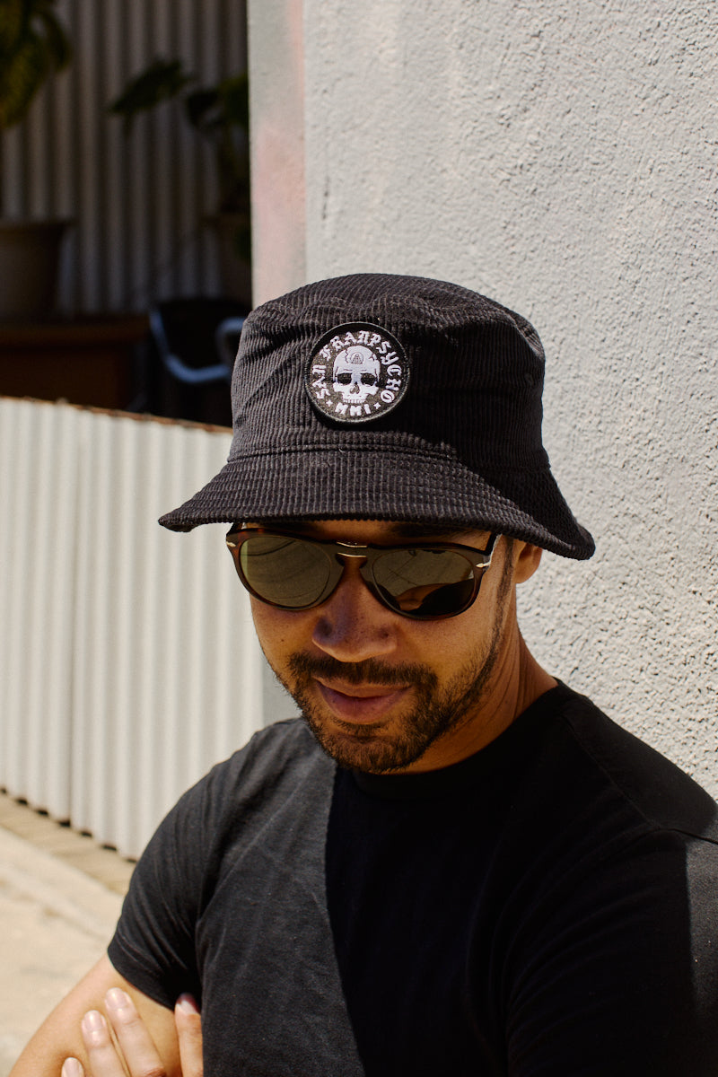 A man wearing sunglasses and the Punk Skull Corduroy Bucket Hat with a circular patch stands outside by a textured wall.