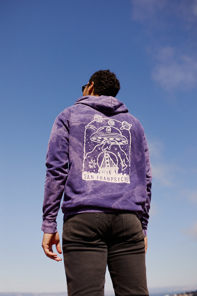 A person wearing the Psycho Invader Pullover Hoodie with a purple and white space-inspired design, hand printed, stands outdoors under a blue sky.