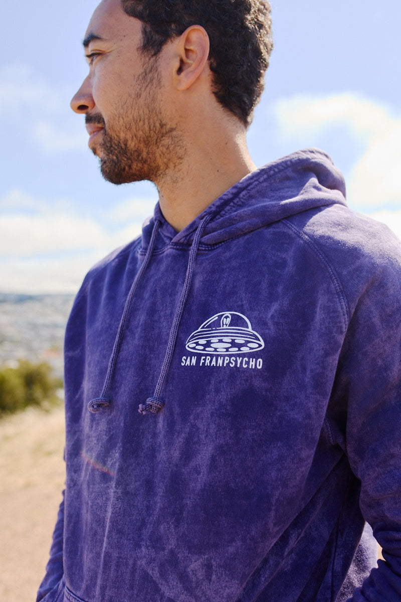 A man stands outdoors looking to the side in a Psycho Invader Pullover Hoodie, hand printed with a UFO design and the San Franpsycho logo.