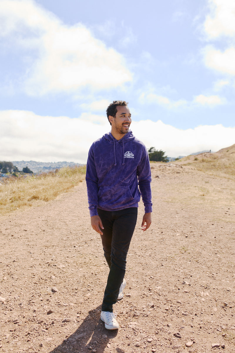 A man wearing a Psycho Invader Pullover Hoodie and black jeans walks along a dirt path beneath a partly cloudy sky.
