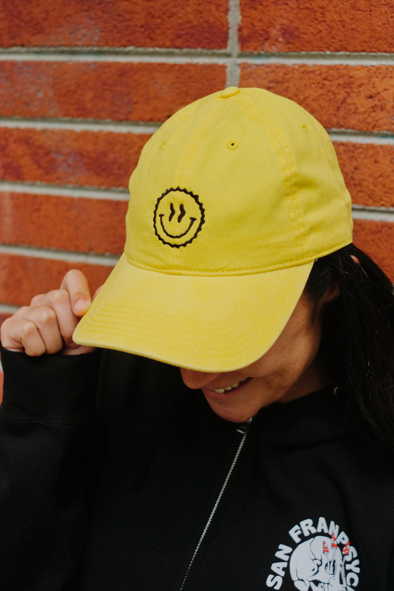 Wearing the Smiley Dad Hat, this person stands before a brick wall, looking down and smiling, enjoying the hat’s comfortable classic fit.