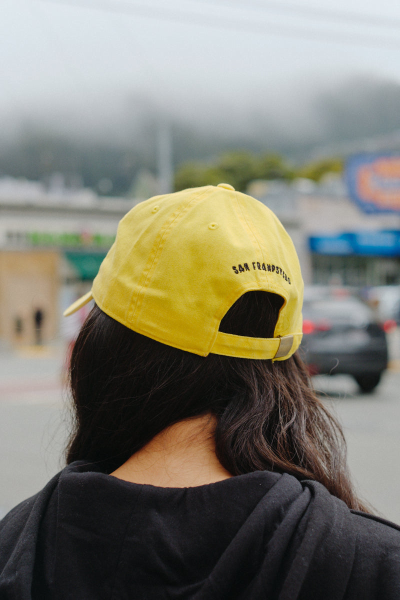 Person with long dark hair wears a classic fit black hoodie and a yellow Smiley Dad Hat, facing away outdoors on a cloudy day.
