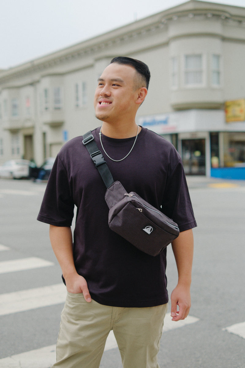 A man in a black t-shirt smiles on a city street with buildings in the background, wearing a Sling Bag featuring an adjustable crossbody strap.