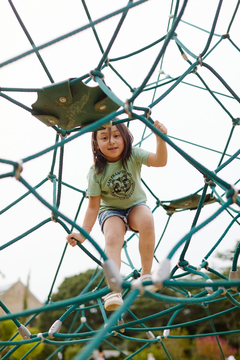 A young child wearing a Bear Essentials Youth Tee climbs on a rope structure at the playground, smiling at the camera.