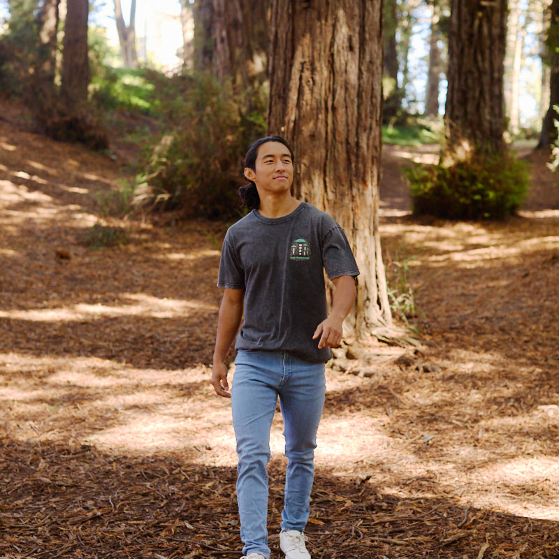 A person strolls through a sunlit forest of tall trees, wearing the Mind Bloom Tee with a skull and mushroom graphic, paired with jeans and white shoes.