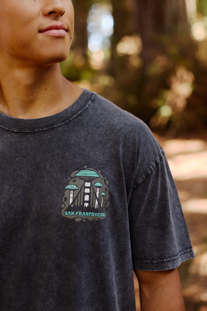 Young person outdoors wearing the Mind Bloom Tee, a mineral wash shirt with a UFO, subtle skull and mushroom graphics, and San Franpsycho text.