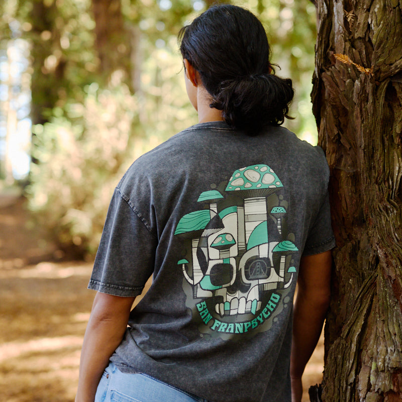 A person in jeans and the Mind Bloom Tee—featuring a skull and mushroom graphic—leans against a tree trunk in a forest, capturing a scene inspired by Erik Abel’s signature style.