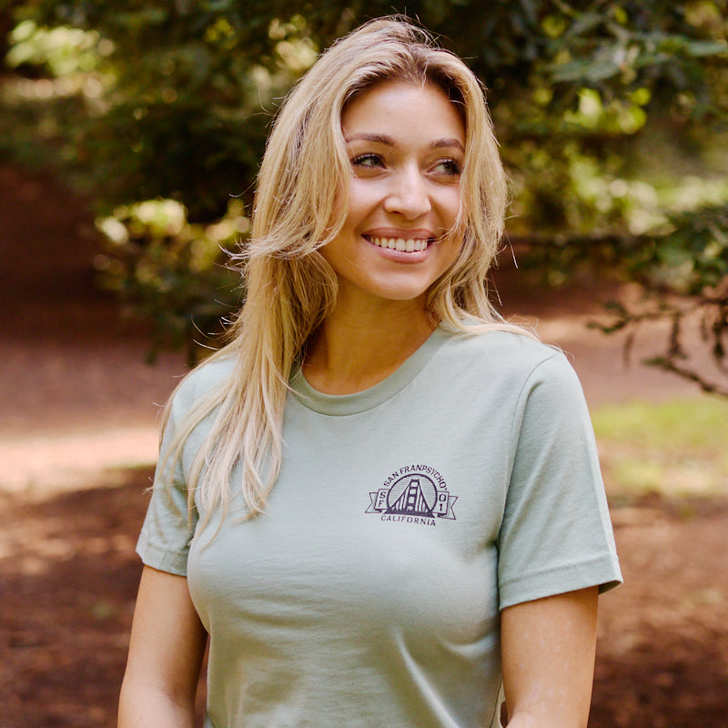 A smiling woman with long blonde hair wears the Bear Essentials Tee in light green, sitting outdoors among trees and capturing a relaxed California bear vibe.