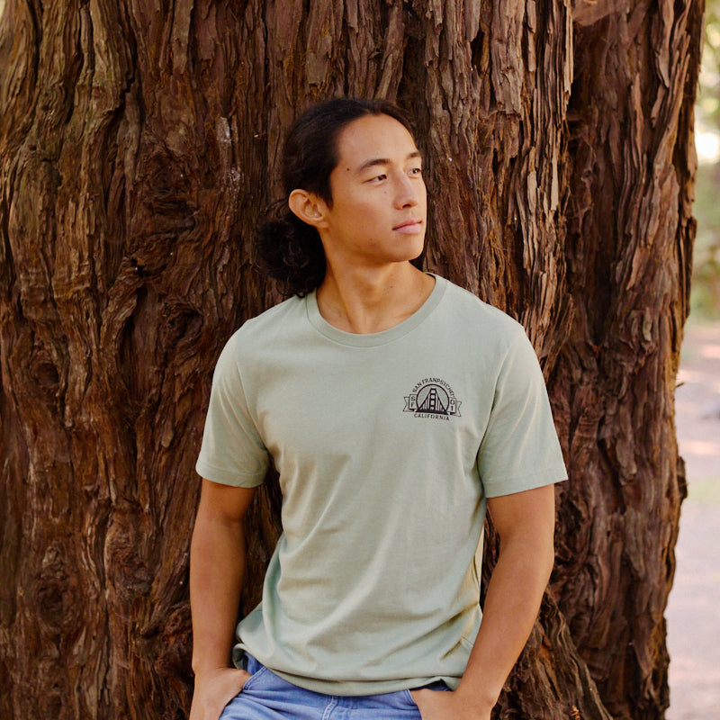 Wearing the Bear Essentials Tee, a man leans against a tree outdoors, gazing to the side and embodying San Francisco's laid-back vibe.