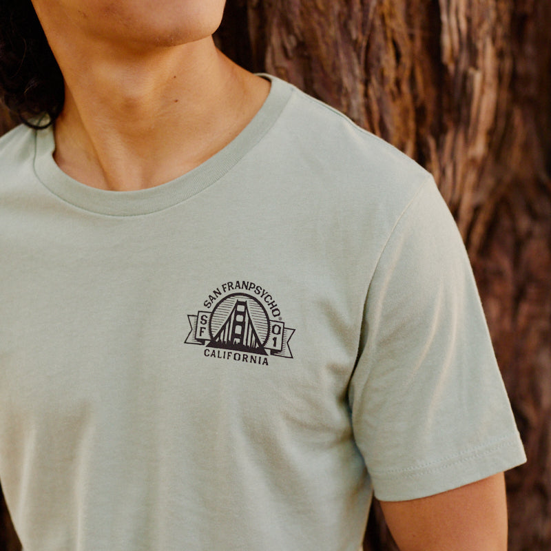 A person wearing the Bear Essentials Tee in light green, featuring a San Francisco, California graphic, stands beside a tree.