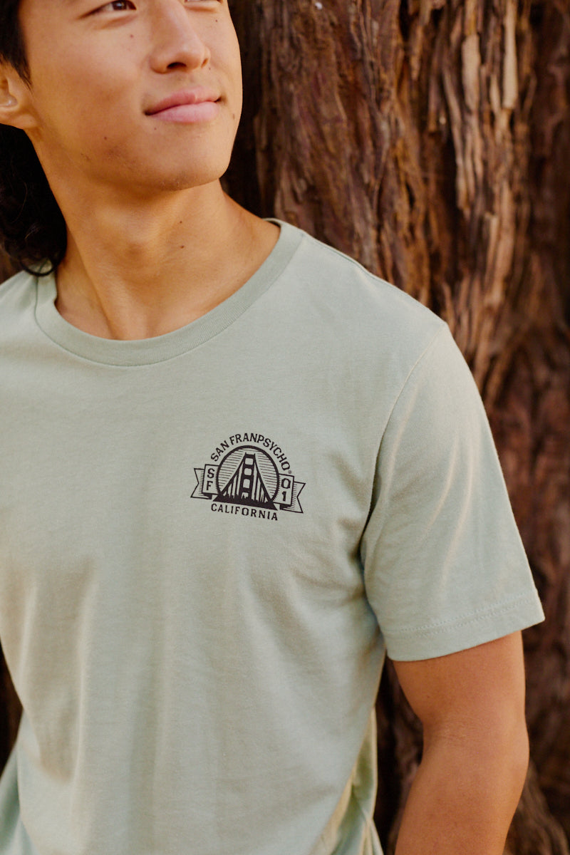 A person wearing the Bear Essentials Tee in light green, featuring a San Francisco, California graphic, stands beside a tree.