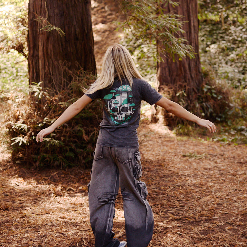 A person with long blond hair and baggy clothes stands with arms outstretched in a forest among tall trees, wearing the Mind Bloom Tee—a mineral wash shirt featuring a skull and mushroom graphic.