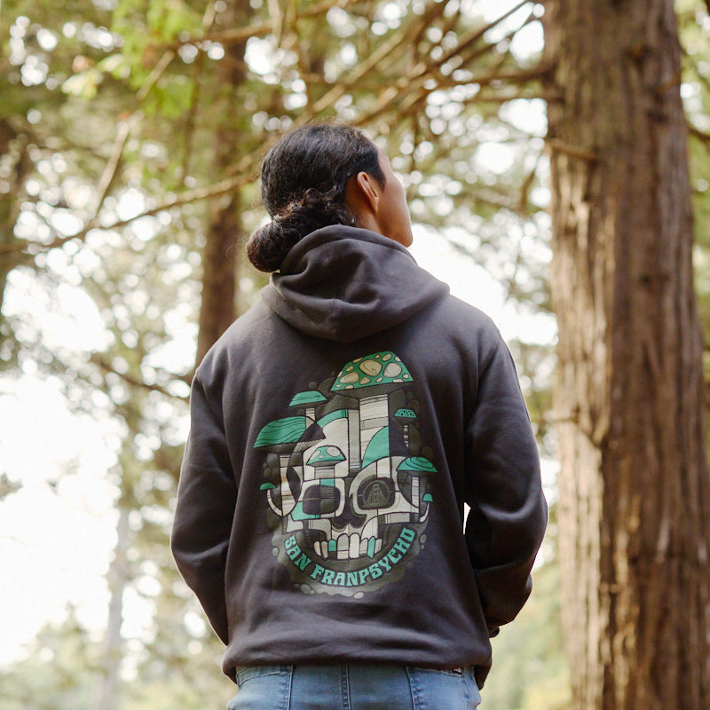 A person wearing the Mind Bloom Pullover Hoodie, with a vibrant skull and mushroom-inspired graphic (in the style of Erik Abel), stands in a forest facing away from the camera, highlighting the hoodie’s unique artistic flair.
