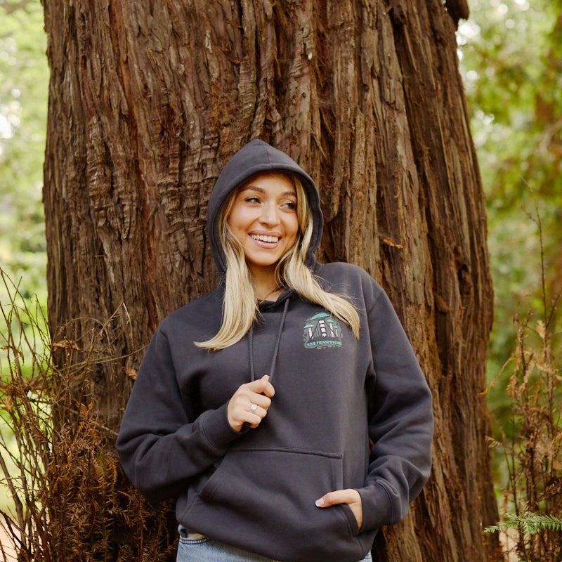 A smiling woman in the Mind Bloom Pullover Hoodie stands against a large tree trunk in a forest.