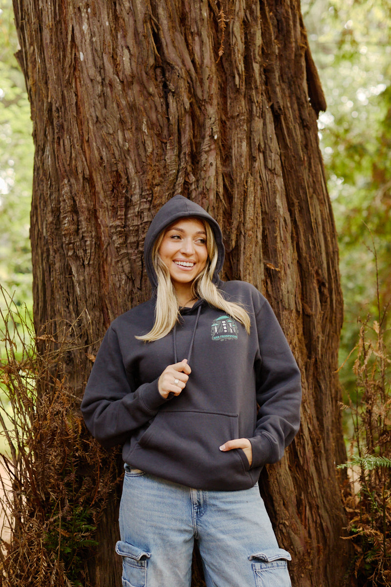 A smiling woman in the Mind Bloom Pullover Hoodie stands against a large tree trunk in a forest.