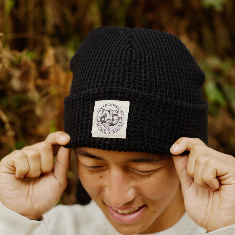 A smiling person in a cream sweater adjusts a Bear Essentials Black Beanie with a patch while standing outdoors.