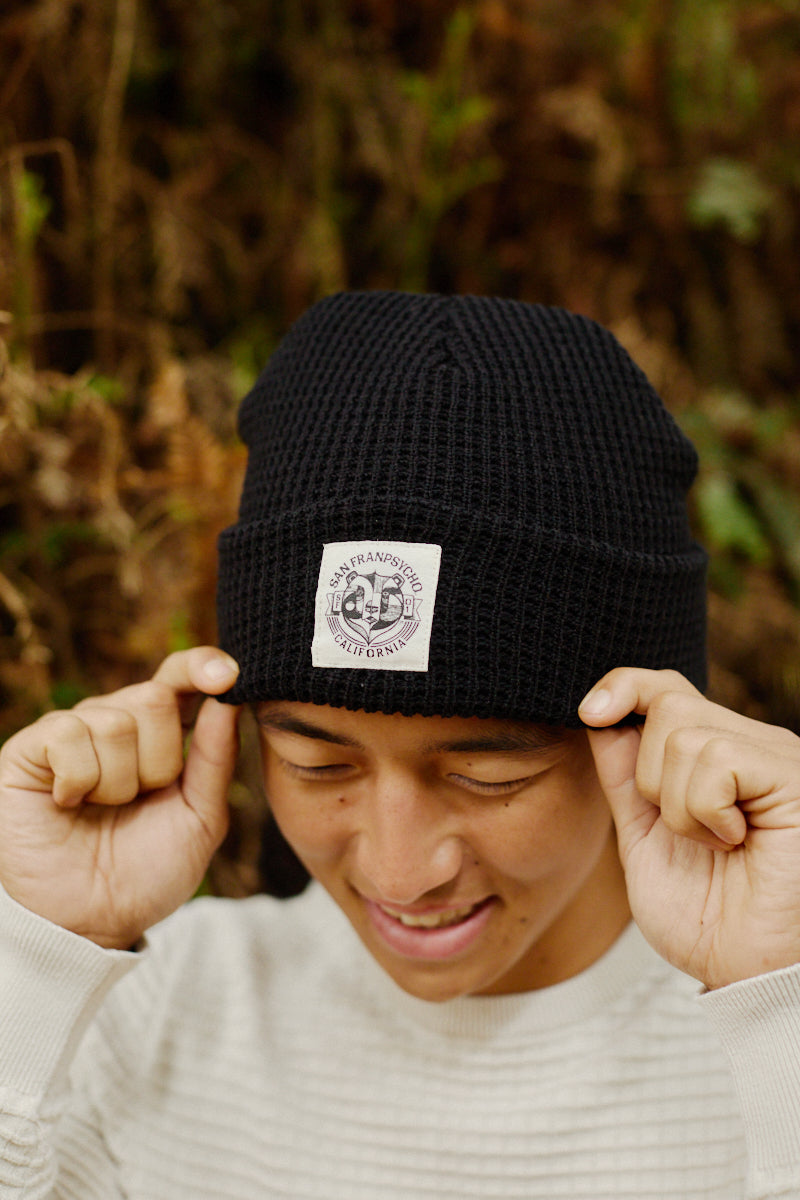 A smiling person in a cream sweater adjusts a Bear Essentials Black Beanie with a patch while standing outdoors.