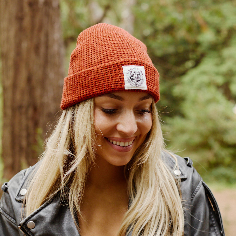 Woman with long blonde hair wearing a Bear Essentials Rust Beanie and black leather jacket, smiling outdoors in a forest.