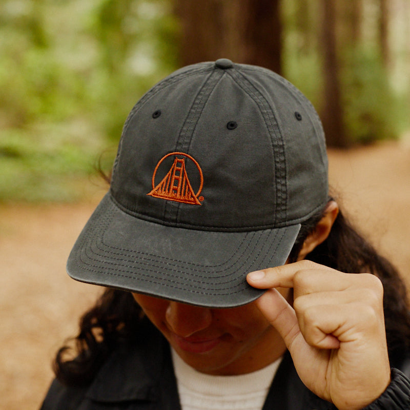 A person in a forest touches the brim of a faded black Rust Logo Dad Hat, which features an orange Golden Gate Bridge design.
