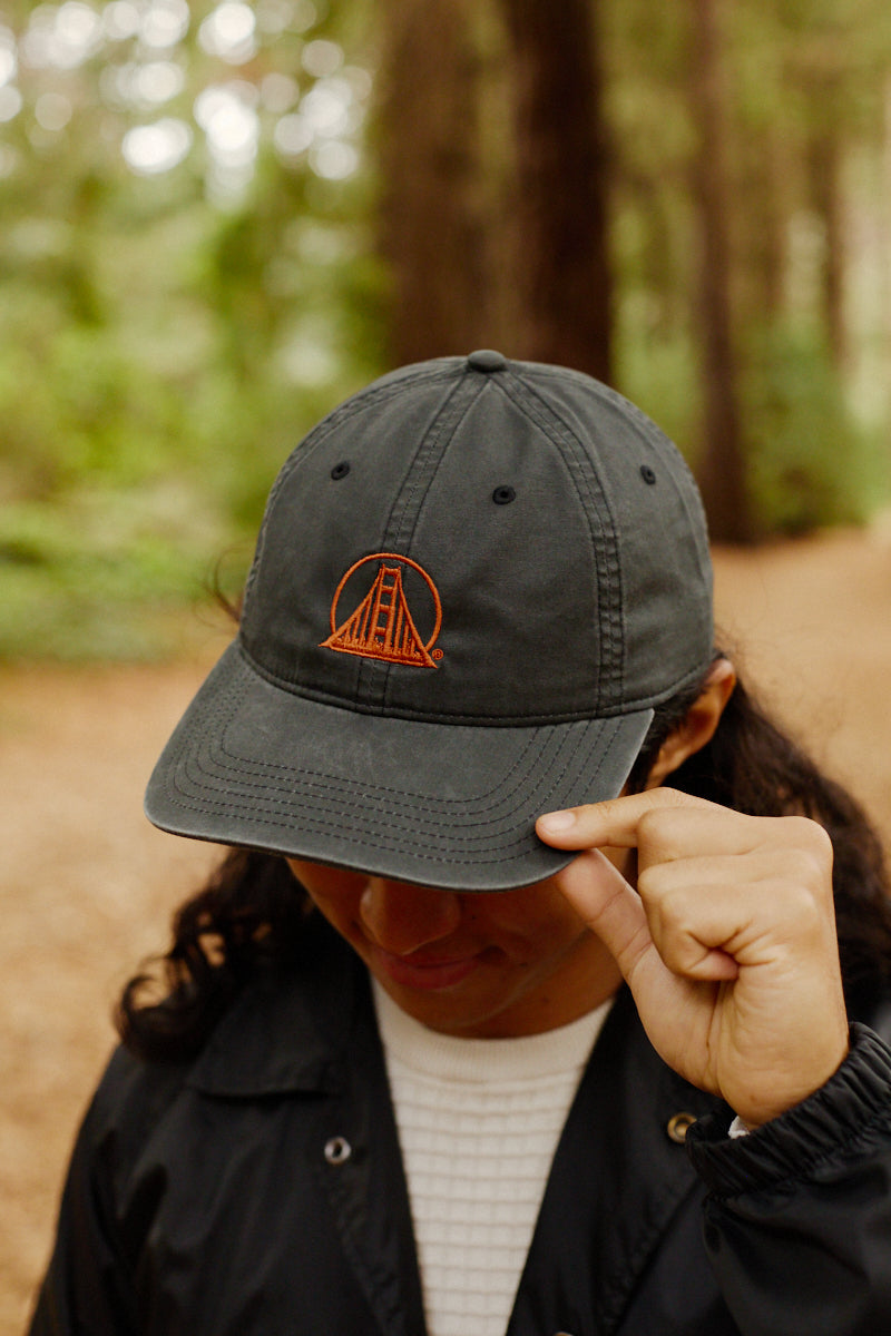 A person in a forest touches the brim of a faded black Rust Logo Dad Hat, which features an orange Golden Gate Bridge design.